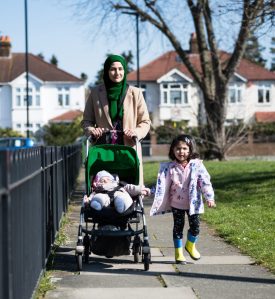 Full length front view of early 30s mother, 2 year old daughter, and 5 month old son in stroller approaching camera on late winter walk in West London suburb.