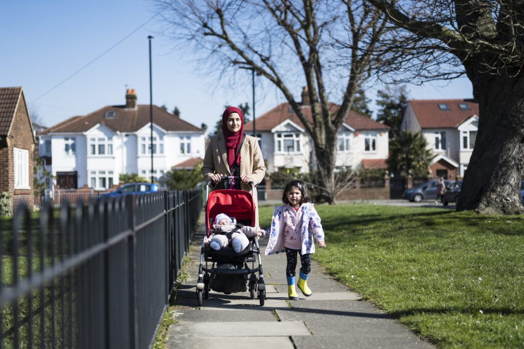Full length front view of early 30s mother, 2 year old daughter, and 5 month old son in stroller approaching camera on late winter walk in West London suburb.